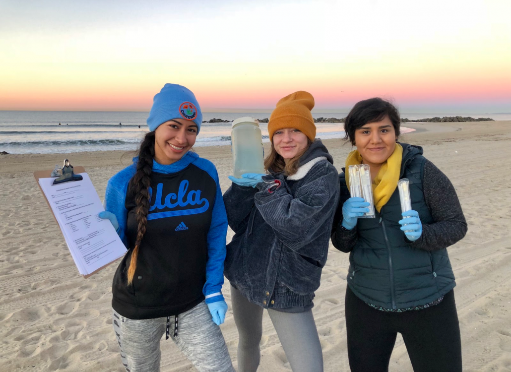 Student researchers on the beach hold up water samples for the camera