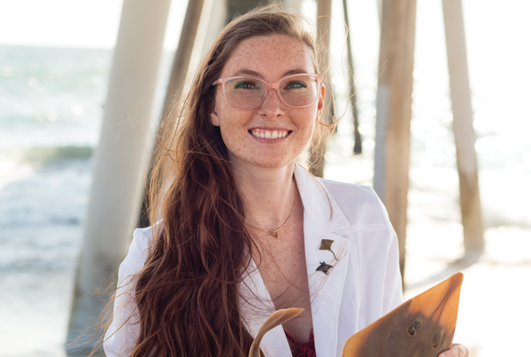 Image of Kelsi Rutledge on a beach in a lab coat, holding a preserved museum specimen of ray.