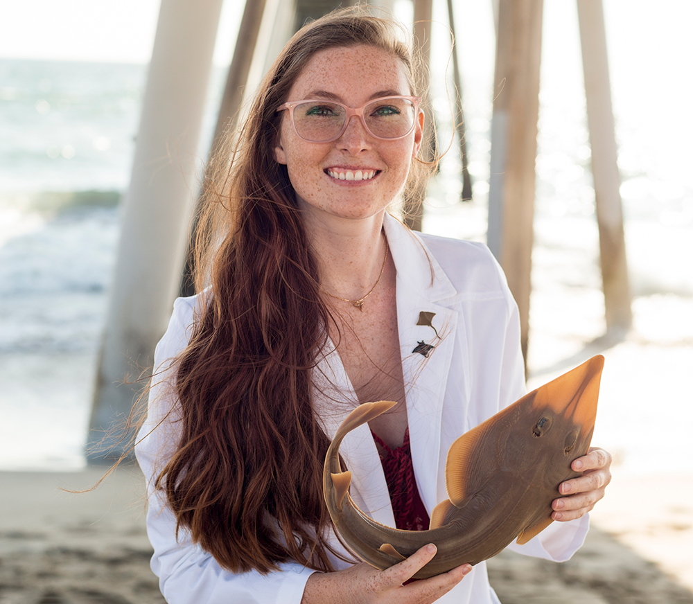 Image of Kelsi Rutledge on a beach in a lab coat, holding a preserved museum specimen of ray.