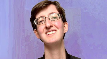 Photo of Christopher, a white genderqueer man with short brown hair, small-framed glasses, a white t-shirt, and a black bomber jacket with a trans pride pin, smiles to the camera in front of a lavender background.
