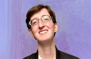 Photo of Christopher, a white genderqueer man with short brown hair, small-framed glasses, a white t-shirt, and a black bomber jacket with a trans pride pin, smiles to the camera in front of a lavender background.