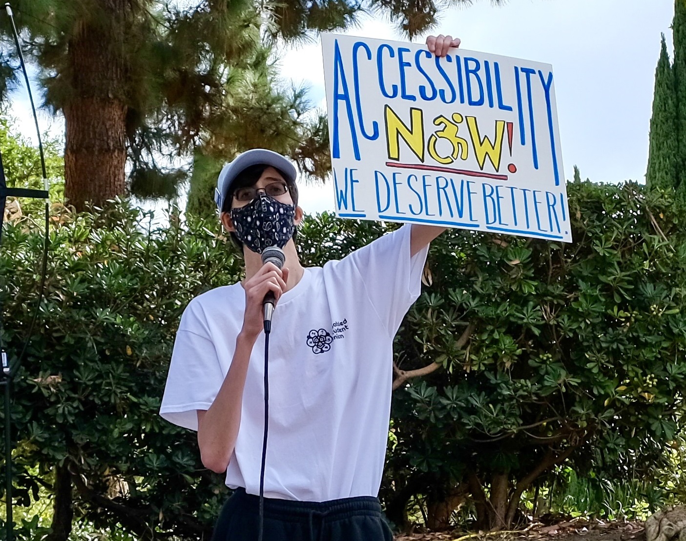 Photo of Christopher, a tall, thin, white genderqueer man with short brown hair tucked under a sky blue baseball cap, small-framed glasses, a black space-themed mask, a white Disabled Student Union t-shirt, black sweatpants, and purple shoes, standing in front of foliage beside Kaplan Hall at UCLA. He speaks into a microphone and holds up a protest sign reading “Accessibility Now! We Deserve Better!” in blue and yellow lettering. Beside him on the ground sits a teal water bottle with stickers on it. 
