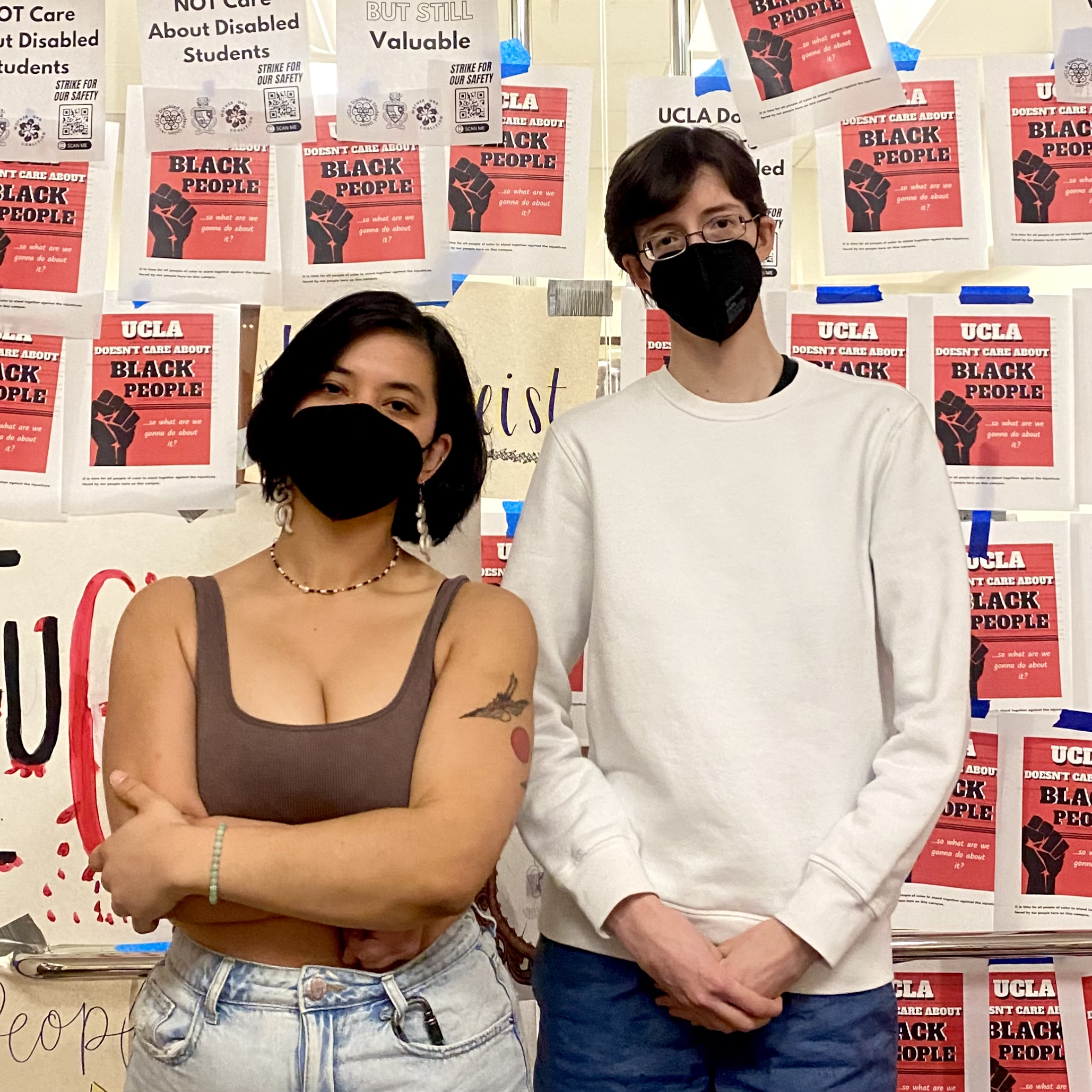 Photo of Quinn (left) and Christopher (right) standing with stern expressions in front of the UCLA Chancellor’s Office doors plastered with flyers and posters with anti-racist and pro-disabled messages. Quinn is a Brown genderfluid person with chin length dark hair, a black KN95 mask, a gold necklace and matching dangly earrings, a rose tattoo, and a brown tank top tucked into light-wash jeans. Christopher is a white genderqueer man with short brown hair, small-framed glasses, a black KN95 mask, a white crewneck sweater, and blue shorts. 