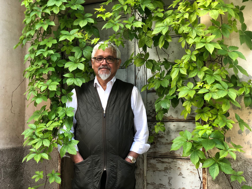 Author Amitav Ghosh in a white long sleeve collar shirt with a black vest standing outside, with green overgrown vines hanging all around him. 