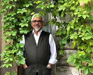 Author Amitav Ghosh in a white long sleeve collar shirt with a black vest standing outside, with green overgrown vines hanging all around him.
