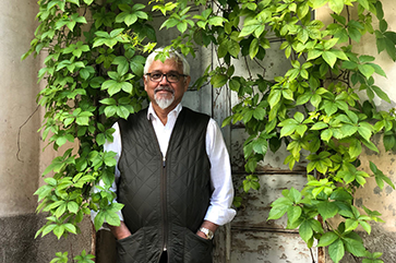 Author Amitav Ghosh in a white long sleeve collar shirt with a black vest standing outside, with green overgrown vines hanging all around him.