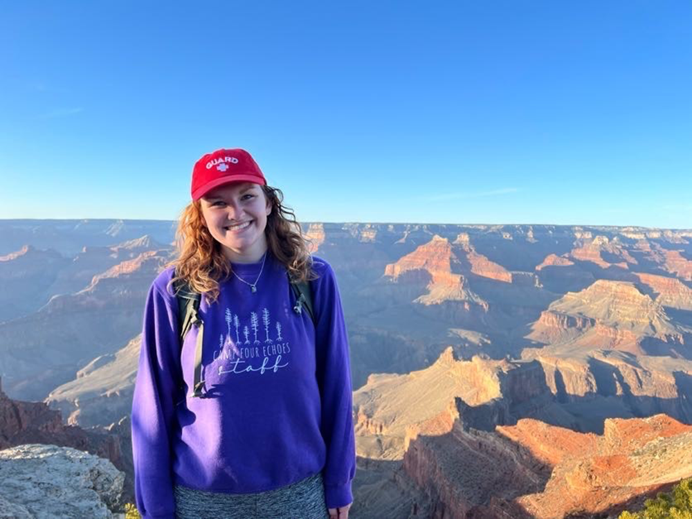 Gillian Smith smiling while wearing a purple sweater and red baseball hat, with canyon formations and a blue afternoon sky in the background. 