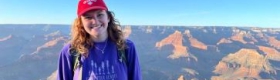 Gillian Smith smiling while wearing a purple sweater and red baseball hat, with canyon formations and a blue afternoon sky in the background.