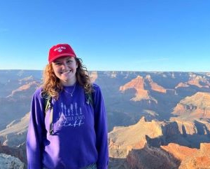 Gillian Smith smiling while wearing a purple sweater and red baseball hat, with canyon formations and a blue afternoon sky in the background.