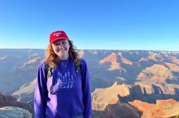 Gillian Smith smiling while wearing a purple sweater and red baseball hat, with canyon formations and a blue afternoon sky in the background.