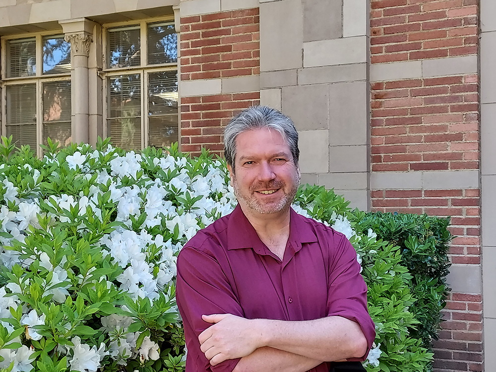 Michael Berry smiling in a dark magenta dress shirt, arms crossed before him, with a green bush blooming white flowers and a brick building with windows in the background. 