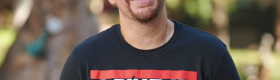 Adam Bradley smiling, both hands in his pockets, in a black short-sleeved shirt that reads: "a beat at its best is a reason to rhyme," with trees and plants in the background.