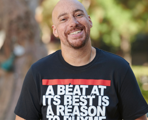 Adam Bradley smiling, both hands in his pockets, in a black short-sleeved shirt that reads: "a beat at its best is a reason to rhyme," with trees and plants in the background.