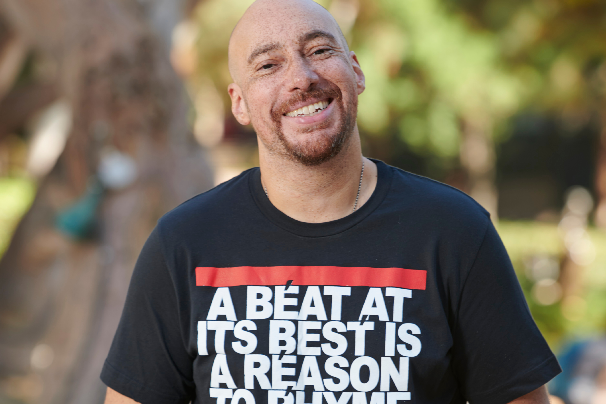 Adam Bradley smiling, both hands in his pockets, in a black short-sleeved shirt that reads: "a beat at its best is a reason to rhyme," with trees and plants in the background.