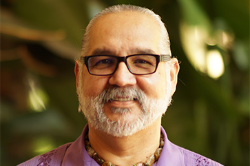 Alfred Herrera in a purple guayabera shirt with a blurred outdoor landscape in the background.