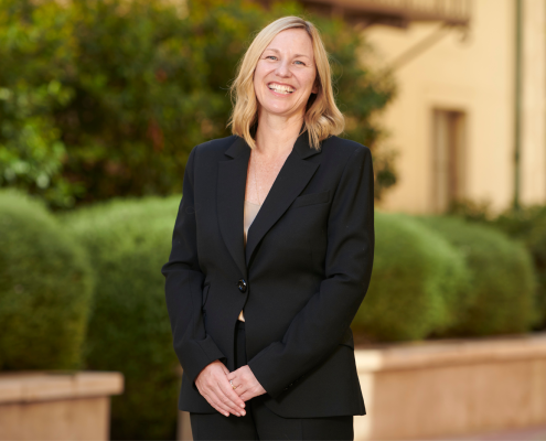 Amander Clark smiling in a black two-piece suit, hands clasped before her, with shrubs inside stone planters in the background