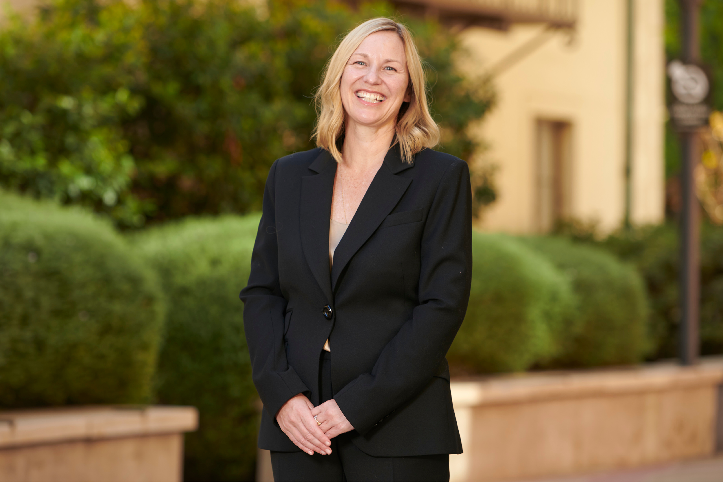 Amander Clark smiling in a black two-piece suit, hands clasped before her, with shrubs inside stone planters in the background