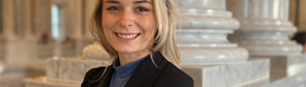 Ariella Gaughan smiling in a black sport coat, with marble columns in the background.