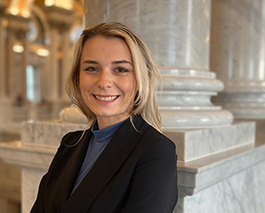 Ariella_363 Ariella Gaughan smiling in a black sport coat, with marble columns in the background.