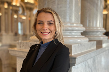 Ariella Gaughan smiling in a black sport coat, with marble columns in the background.