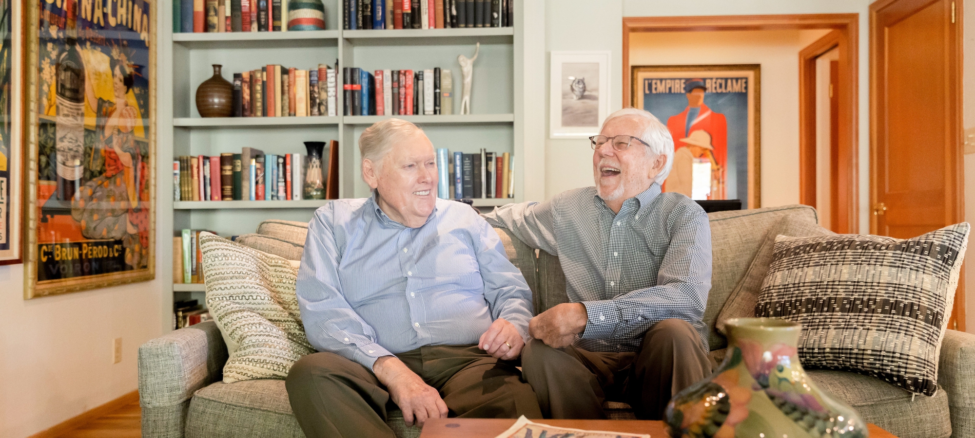 David Bohne (left) smiling and Tom Bye (right) laughing in business casual attire as they sit on a beige couch with plenty of pillows, with a coffee table and an empty flower vase in the forefront, and a bookcase, hung photographs and posters in the background.