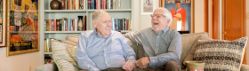 David Bohne (left) smiling and Tom Bye (right) laughing in business casual attire as they sit on a beige couch with plenty of pillows, with a coffee table and an empty flower vase in the forefront, and a bookcase, hung photographs and posters in the background.
