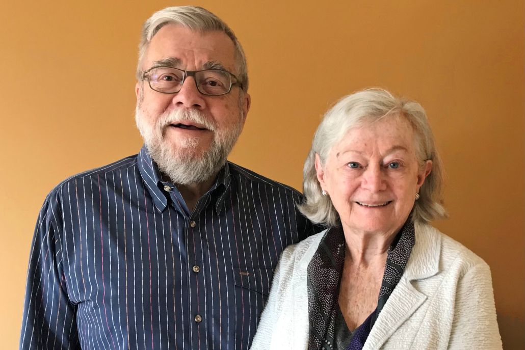 Michael Casteel (left) wearing glasses and in a blue dress shirt with red, green, and yellow stripes, and Cathy Casteel (right) in a white linen coat, both smiling, against a light orange background.