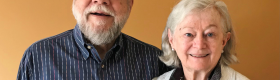 Michael Casteel (left) wearing glasses and in a blue dress shirt with red, green, and yellow stripes, and Cathy Casteel (right) in a white linen coat, both smiling, against a light orange background.