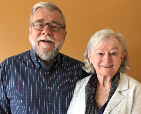 Michael Casteel (left) wearing glasses and in a blue dress shirt with red, green, and yellow stripes, and Cathy Casteel (right) in a white linen coat, both smiling, against a light orange background.