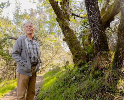 Hollis Lenderking on a hiking trail wearing hiking boots, khaki pants, and a collared shirt and grey sweater, both hands inside his sweater pockets, surrounded by trees and a blue sky in the background.