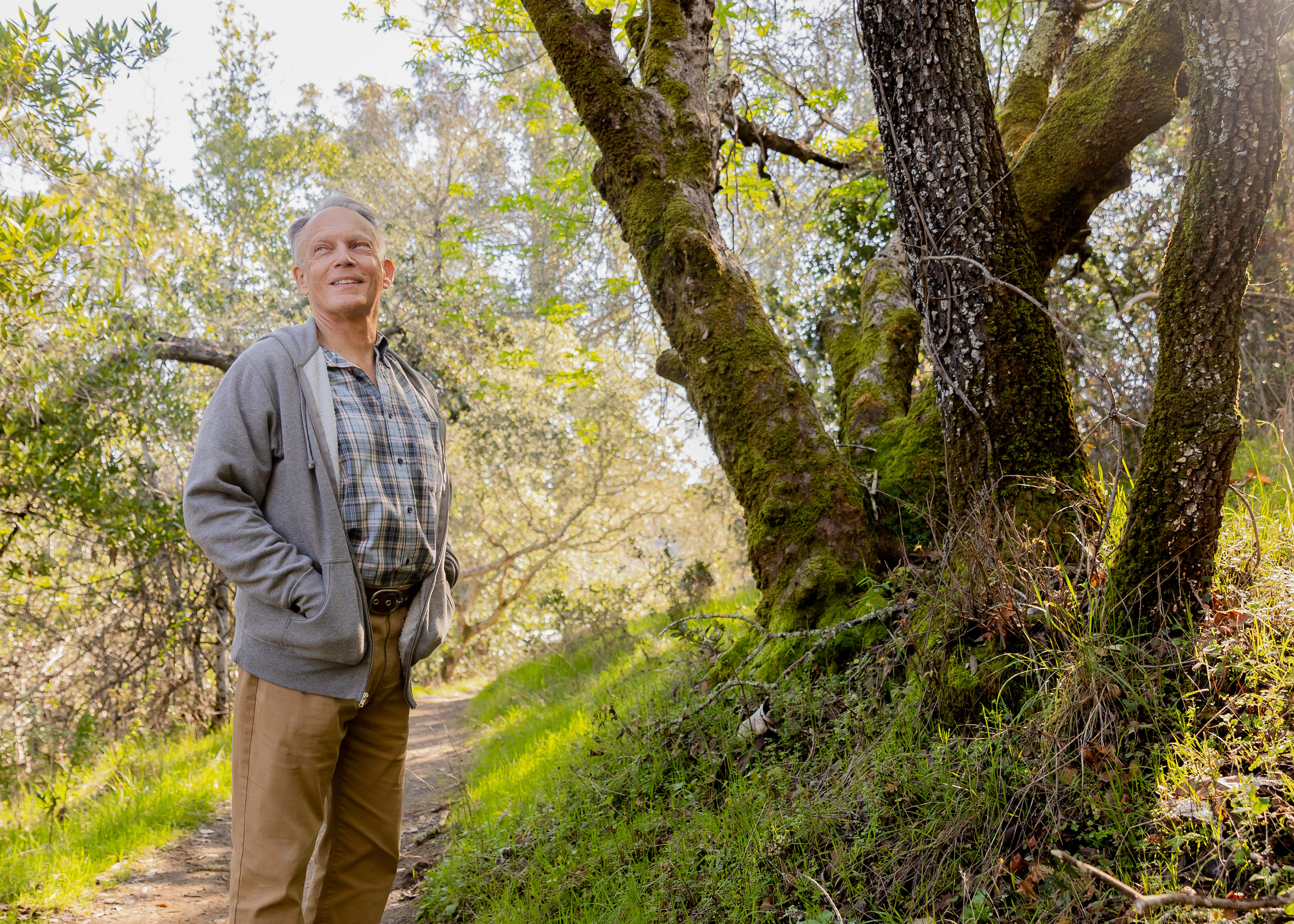 Hollis Lenderking on a hiking trail wearing hiking boots, khaki pants, and a collared shirt and grey sweater, both hands inside his sweater pockets, surrounded by trees and a blue sky in the background.