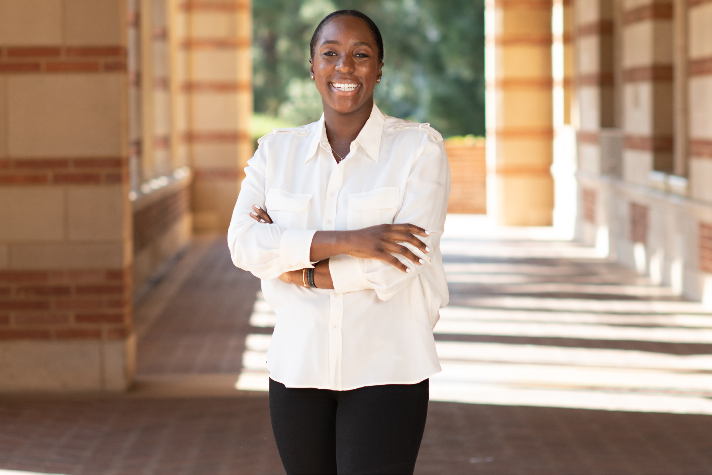 Jessica Lee, smiling with arms folded in a white-collared shirt and black pants, is standing in a sunlit walkway under the Royce Hall portico with trees in the background.