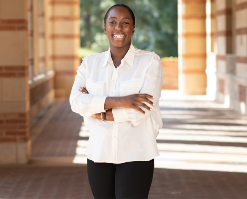 Jessica Lee, smiling with arms folded in a white-collared shirt and black pants and boots, is standing in a sunlit walkway under the Royce Hall portico with trees in the background.