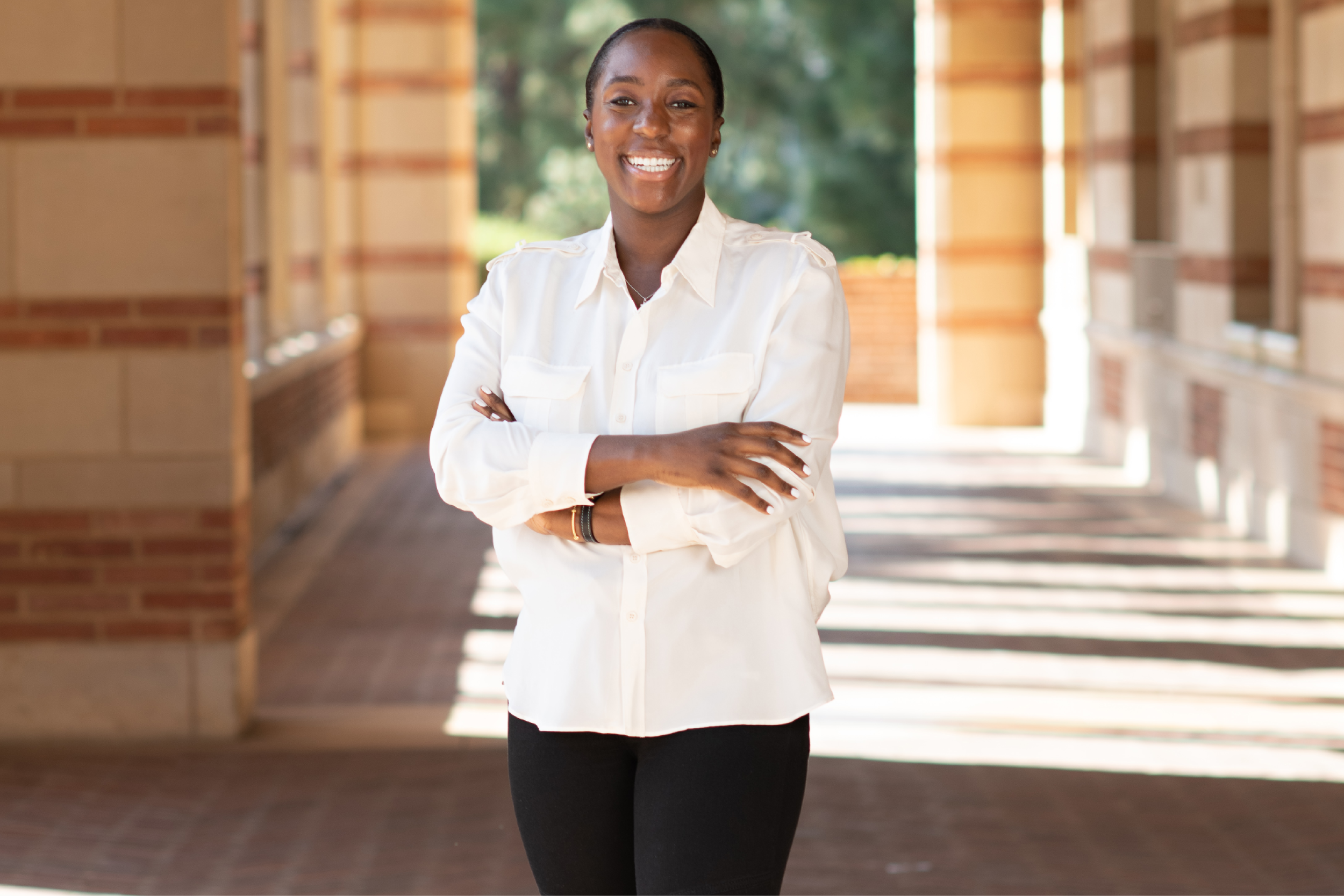 Jessica Lee, smiling with arms folded in a white-collared shirt and black pants and boots, is standing in a sunlit walkway under the Royce Hall portico with trees in the background.