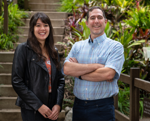 Jordyn Moscoso (left) smiling in a black leather jacket and orange floral shirt, hands clasped before her; Jon Aurnou (right) smiling in a blue checkered short-sleeve collar shirt, arms crossed; concrete steps and green plants in the background.