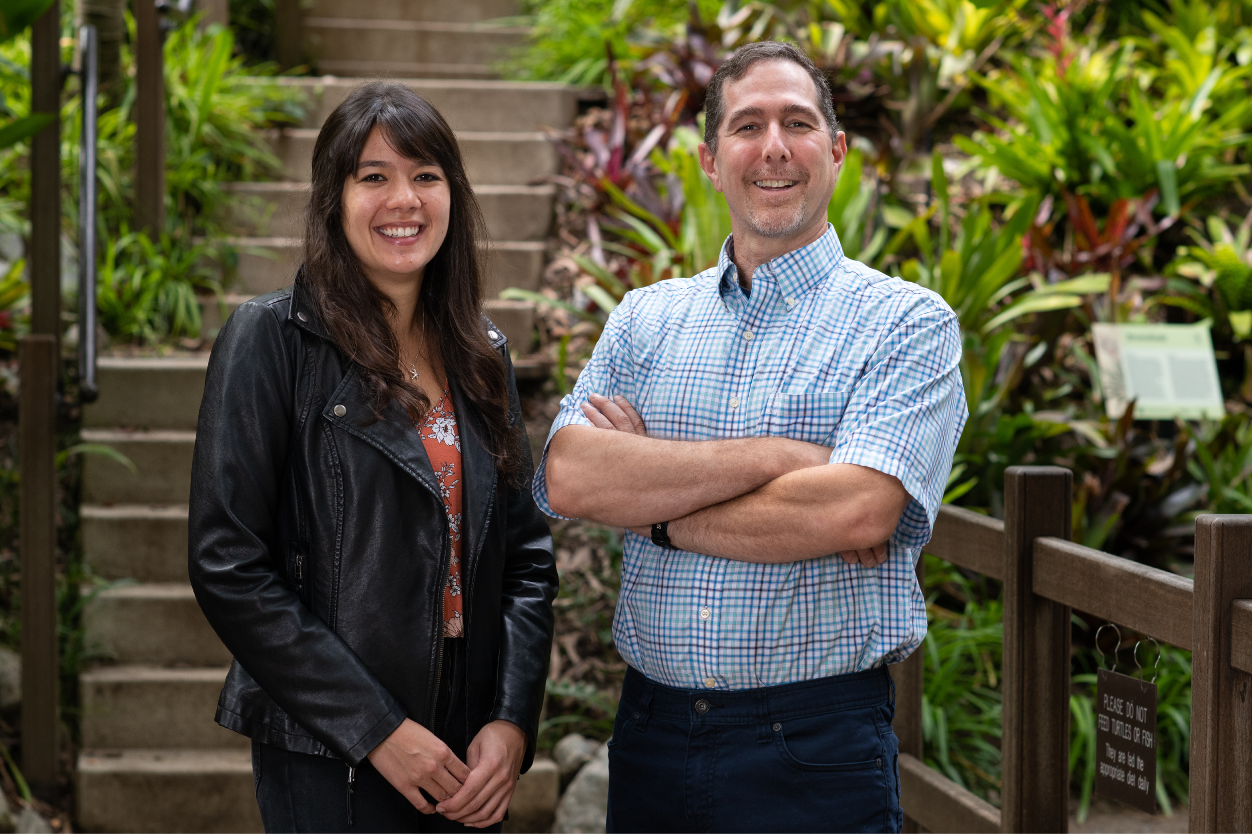 Jordyn Moscoso (left) smiling in a black leather jacket and orange floral shirt, hands clasped before her; Jon Aurnou (right) smiling in a blue checkered short-sleeve collar shirt, arms crossed; concrete steps and green plants in the background.