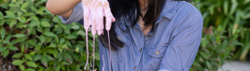 Laurie Tan in a blue jean button-up shirt with sleeves rolled up, sitting on grass and smiling as she plays with a pink, gooey substance, green plants in the background