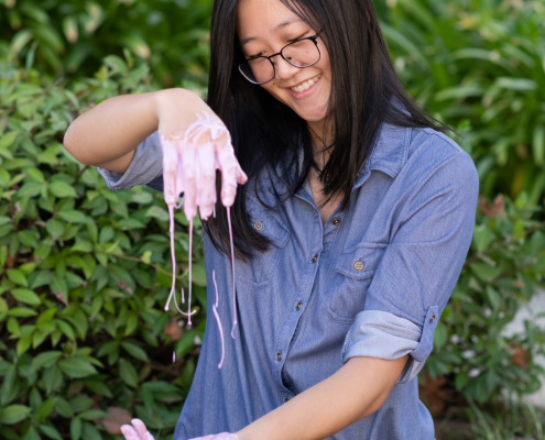 Laurie Tan in a blue jean button-up shirt with sleeves rolled up, sitting on grass and smiling as she plays with a pink, gooey substance, green plants in the background