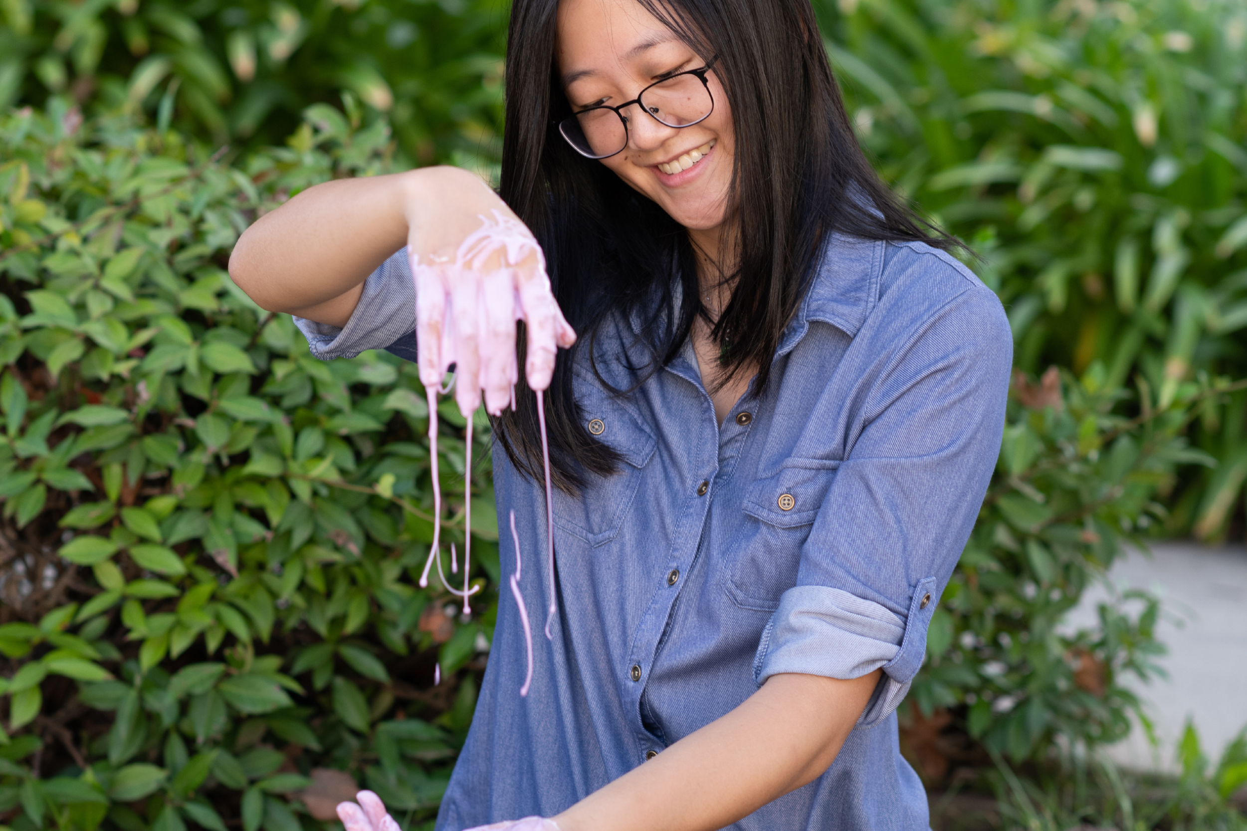 Laurie Tan in a blue jean button-up shirt with sleeves rolled up, sitting on grass and smiling as she plays with a pink, gooey substance, green plants in the background