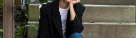 Makena Tinney in a black blazer and blue jeans, left hand on her chin as she sits at the base of concrete steps lined with a metallic railing and green plants.