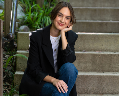 Makena Tinney in a black blazer and blue jeans, left hand on her chin as she sits at the base of concrete steps lined with a metallic railing and green plants.
