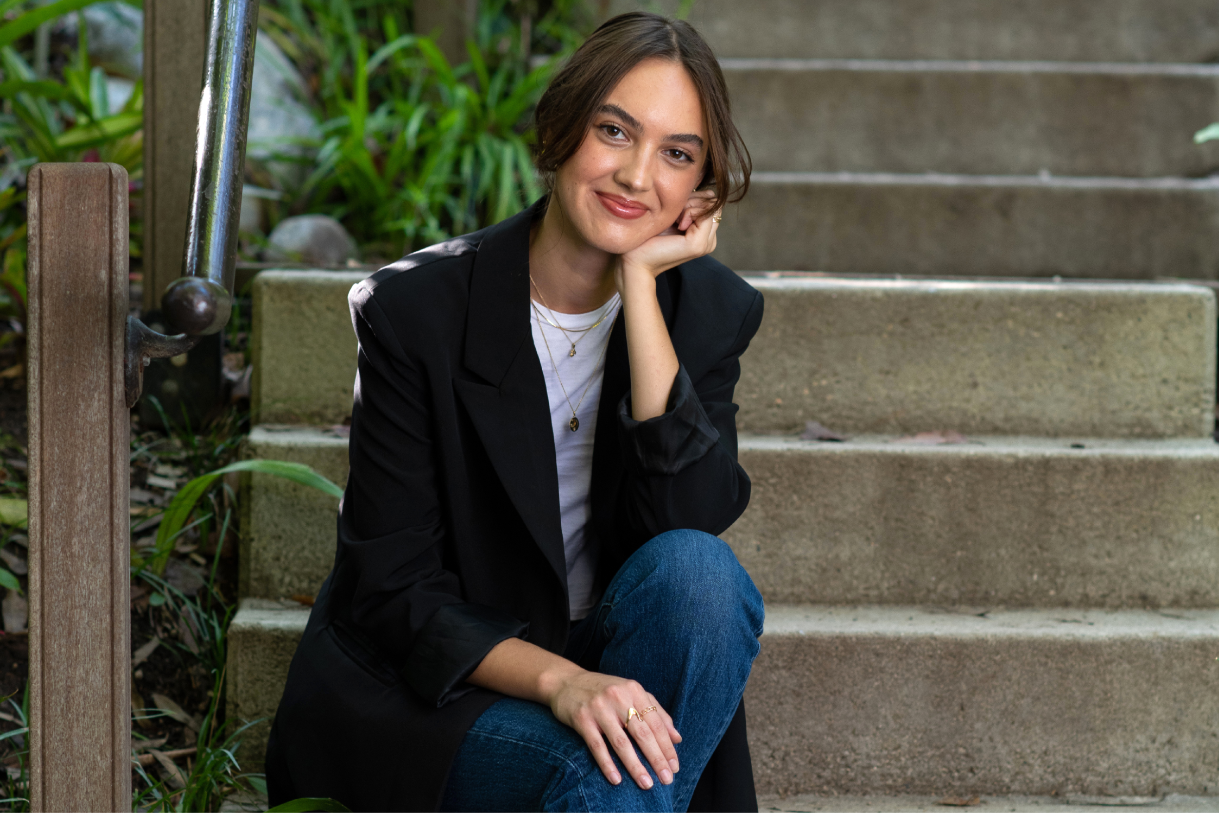 Makena Tinney in a black blazer and blue jeans, left hand on her chin as she sits at the base of concrete steps lined with a metallic railing and green plants.