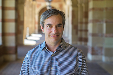 Michael Rothberg in a blue checkered dress shirt, with a walkway lined with stone columns in the background.