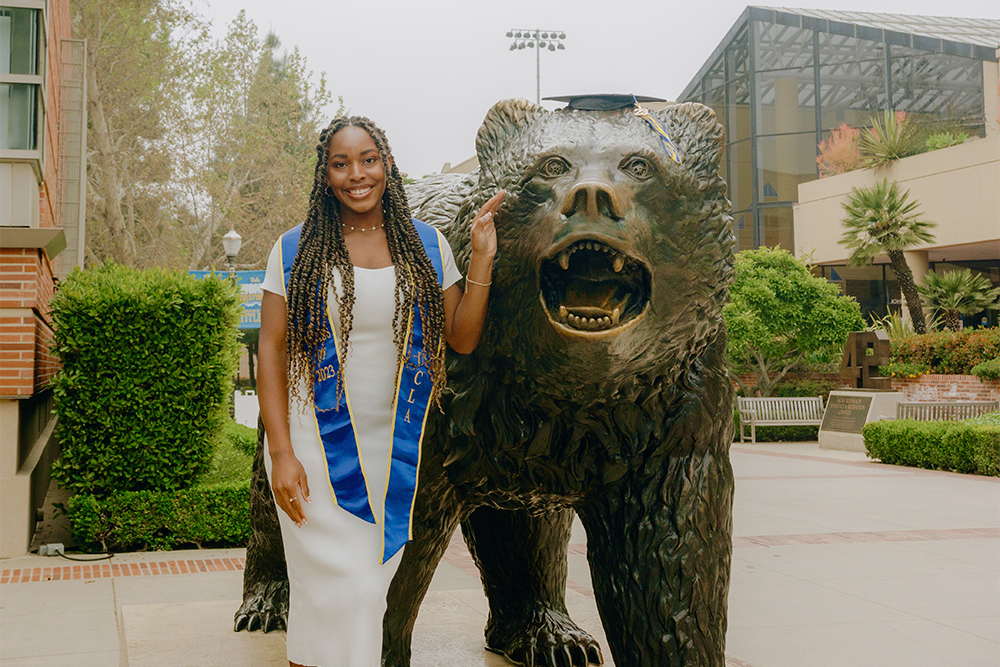 Nene Usim in a white dress and blue graduation sash, posing next to a bronze bruin bear statue