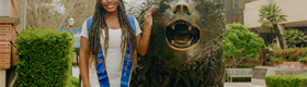 Nene Usim in a white dress and blue graduation sash, posing next to a bronze bruin bear statue