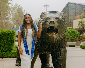 Nene_363 Nene Usim in a white dress and blue graduation sash, posing next to a bronze bruin bear statue