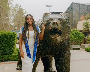 Nene Usim in a white dress and blue graduation sash, posing next to a bronze bruin bear statue