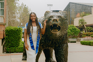 Nene Usim in a white dress and blue graduation sash, posing next to a bronze bruin bear statue