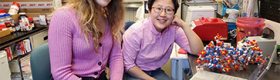 UCLA Graduate Student Noelle Mitchell wearing a pink sweater (left), and Dr. Kyungae Yang of Columbia in a pink button-up shirt (right), smiling in a research lab with equipment all around them.