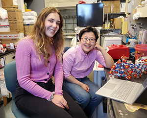UCLA Graduate Student Noelle Mitchell wearing a pink sweater (left), and Dr. Kyungae Yang of Columbia in a pink button-up shirt (right), smiling in a research lab with equipment all around them.